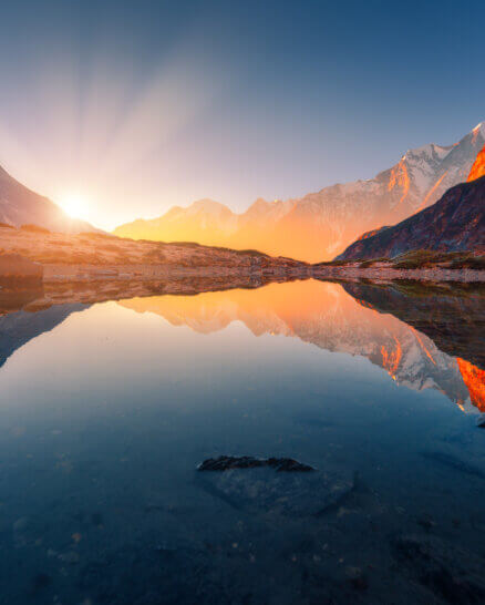 Beautiful landscape with high mountains with illuminated peaks, stones in mountain lake, reflection, blue sky and yellow sunlight in sunrise. Nepal. Amazing scene with Himalayan mountains. Himalayas